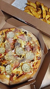 Pizza with ham, white sausages and fried potatoes in cardboard boxes