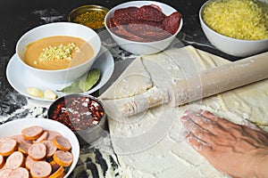 Pizza ingredients. hand rolls out the dough. rolling pin and flour. sliced salami and sausages. grated cheese and pizza sauce