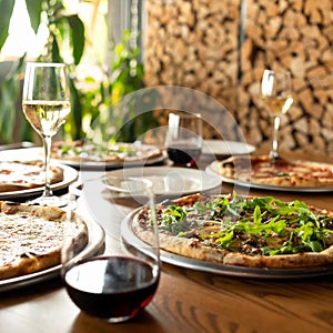 Pizza party. Assorted five different pizzas on wooden restaurant table with glasses of wine on it. Woodpile of firewood