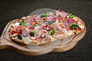 Pizza with salami, ham, vienna sausages, kalamata, olives, broccoli, pelati, pesto. Roman pizza rectangular on wood background