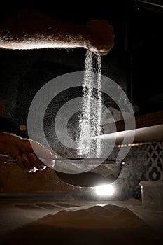 Pizzaiolo& x27;s hands sift flour through a sieve before preparing pizza in poor lighting