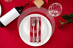 Place setting for Valentines day. Fork and knife on red napkin on plate, wine bottle, glasses, rose on red background