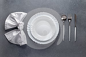 Place setting with white napkin, empty plate and silver cutlery on a gray concrete grunge table. Top view. Festive dinner in a