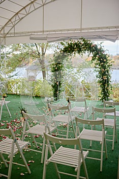 Place for wedding ceremony in tent outdoors, copy space. Wedding arch decorated with flowers and chairs on each side of archway.