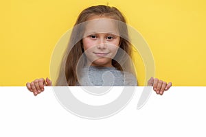 Place for your advertisement. Baby girl pointing to blank white billboard on yellow background, copy space. Emotions