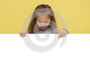 Place for your advertisement. Baby girl pointing to blank white billboard on yellow background, copy space. Emotions