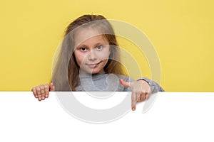 Place for your advertisement. Baby girl pointing to blank white billboard on yellow background  copy space. Emotions
