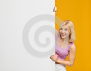 Place for your promo. Joyful woman standing behind white empty board for advertisement over yellow background