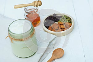 Plain yoghurt in white bowl, honey, cereal, raisin and wood spoon on white wooden table background