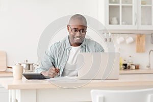 Plan finances. Positive african american mature man calculating taxes and using laptop computer, sitting in kitchen