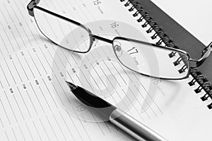 Plan for the week. Glasses in a metal frame and pen with an open gold pen on the organizer. Black and white photography