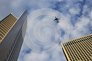The plane flies over high-rise modern buildings. On the background blue sky with clouds