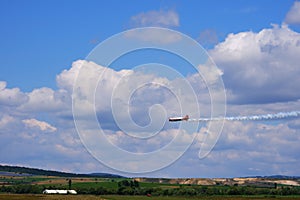 Plane flying with smoke in the sky with clouds over green fields