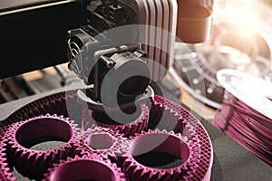 planetary gear machine part is produced by a 3D-printer from pink plastic. filament roll and wooden desk area in background.