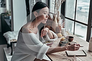 Planning the day. Attractive young woman eating healthy breakfast and writing something down while sitting near the window at home