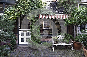 The plant-covered entrance to a small hidden and cozy shop. White door and window frames. An awning protects the window. in front