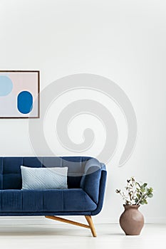 Plant next to navy blue couch with cushion in white living room interior with poster. Real photo