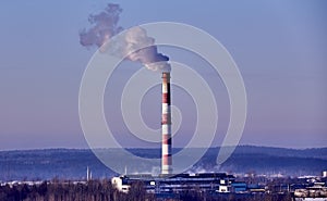 The plant\'s tall brick chimney emits a long cloud of thick white smoke against a blue sky