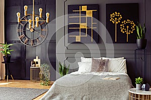 Plants and black and gold posters in grey bedroom interior with clock and bed with pillows. Real photo