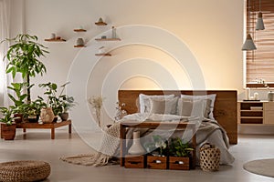 Plants and pouf in dark bedroom interior with light behind wooden bed with headboard. Real photo