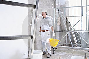 Plasterer man work holds a bucket front the wall of interior construction site wear helmet and protective gloves, ladder and