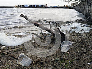 Plastic bottles and glasses on the sandy beach. Dirty water in which zllichny garbage floats. White foam on the shore
