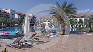 Plastic and fabric umbrellas and lounge chairs are spread out on the tiled pool deck surrounded by buildings with balconies and pa