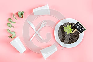 Plastic glasses, forks, knives,plate with earth, a green sprout and a peg stop plastic on a pink.
