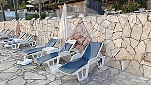 Plastic lounge sunbed with soft mattresses, umbrellas and tables are arranged on a tiled area near the wall. Palm trees grow nearb