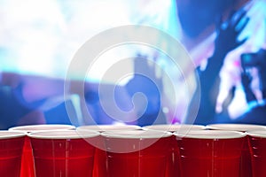 Plastic red party cups in a row in a nightclub full of people dancing on the dance floor in the background.