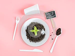 A plastic white plate with black earth, a green sprout, an empty glass, spoons, forks and a peg .