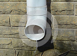 Plastic white rain gutter downspout pipe runs out rain water near house foundation wall. Rainwater flows out of the gutter