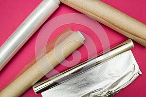 Plastic wrap, aluminum foil and roll of parchment paper on pink background