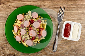 Plate with boiled pasta, fried sausages and tomato sauce, fork, sauce boat with ketchup and mayonnaise on wooden table. Top view
