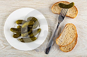 Plate with canned cucumbers, marinated gherkin strung on fork on bread on wooden table. Top view