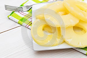 Plate with canned pineapple rings with fork and kitchen towel on white wooden table