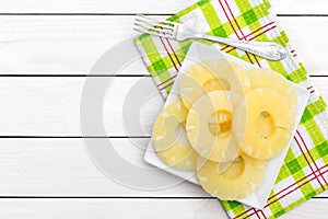 Plate with canned pineapple rings with fork and kitchen towel on white wooden table. Top view. Space for text