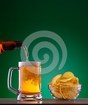 Plate with chips and light beer pouring into mug on green background