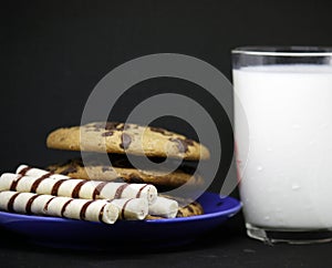 A plate of chocolate chip cookies on a blue plate with a glass of milk on a black background close-up