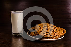 Plate with chocolate chip cookies and a glass of milk on a dark wooden table