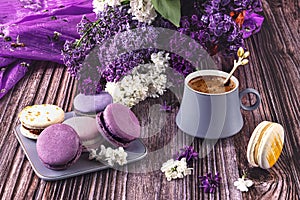 A plate with a colorful French dessert of macaroons on a wooden background and a gray cup of coffee. Purple lilac flowers