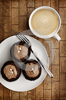 A Plate containing 3 chocolate fairy cakes and a knife and a cup of coffee on a checked wooden background, shot from above