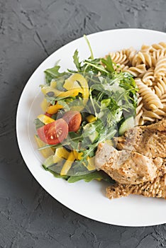 Plate with cooked turkey breast, fresh vegetables salad and whole wheat fusilli pasta on concrete table