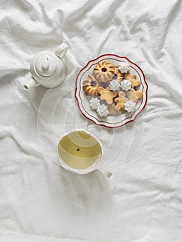 A plate of cookies, a cup of green tea, a teapot on white sheets in the bed, top view