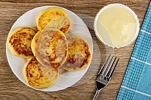 Plate with cottage cheese pancakes, bowl with condensed milk, fork, napkin on wooden table. Top view