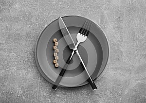 Plate with cutlery and word Anorexia made of wooden cubes on grey table, top view
