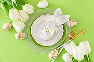 Plate decorated bunny ear napkin, white tulips and Easter eggs on green background. Easter celebration concept. Flat lay. Top view