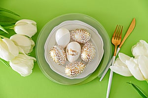 Plate decorated golden Easter eggs and white tulips on green background. Easter celebration concept. Flat lay. Top view