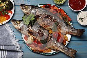 Plate with delicious baked sea bass fish and vegetables on light blue table, flat lay