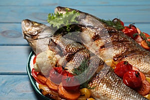 Plate with delicious roasted sea bass fish and vegetables on light blue wooden table, closeup